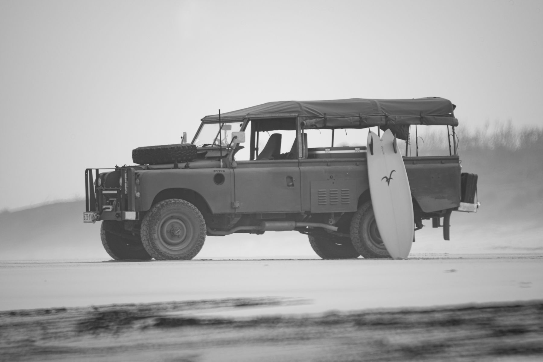 An Aloha surfboard leaning against an old Land Rover on a windswept beach